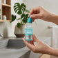 Angle : Prise de vue lifestyle à hauteur d'homme.  Scène : Une main humaine soignée et naturelle tient délicatement le flacon Anua par sa base turquoise, s'apprêtant à l'utiliser. La scène se passe devant une vasque de salle de bain moderne en béton ciré gris. Quelques gouttes d'eau fraîches sont visibles sur les doigts.  Fond : Arrière-plan flou de salle de bain d'architecte avec des plantes d'intérieur (Monstera). (gemini-3.1-flash-image-preview)