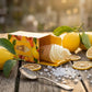 Prompt : Photographie de style food styling haute définition. Le flacon de parfum Arabiyat Sugar "Lemon Sorbet" (tel que vu dans image_8957e6.jpg) glisse doucement hors de sa boîte en carton jaune entrouverte. L'ensemble est posé sur une table de jardin en bois brut, baignée par une lumière dorée de fin d'après-midi. Le flacon en forme de cône de glace à l'italienne est à moitié sorti de l'étui. Tout autour, pour un effet naturel et frais, sont disposés des citrons entiers avec leurs feuilles vertes encore 