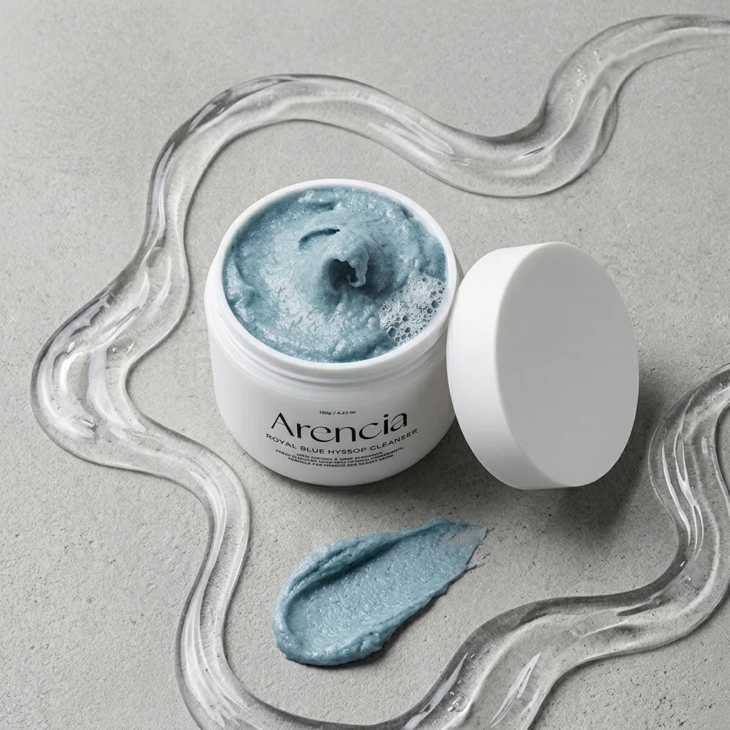 Angle : Nadir parfait (Zénithal à $90^\circ$ - Flat Lay de studio).Scène : Une composition artistique à plat. Le pot de l'image image_cc26e3.jpg est ouvert au centre, dévoilant sa pâte bleue. Son couvercle blanc mat est disposé à côté. Au premier plan, la trace texturée bleue (swatch) est mise en valeur. Tout autour, une traînée sinueuse de gel aqueux transparent et brillant dessine des vagues fluides.Fond & Lumière : Le fond est une plaque de béton ciré gris perle mat. Éclairage de studio très doux et diff