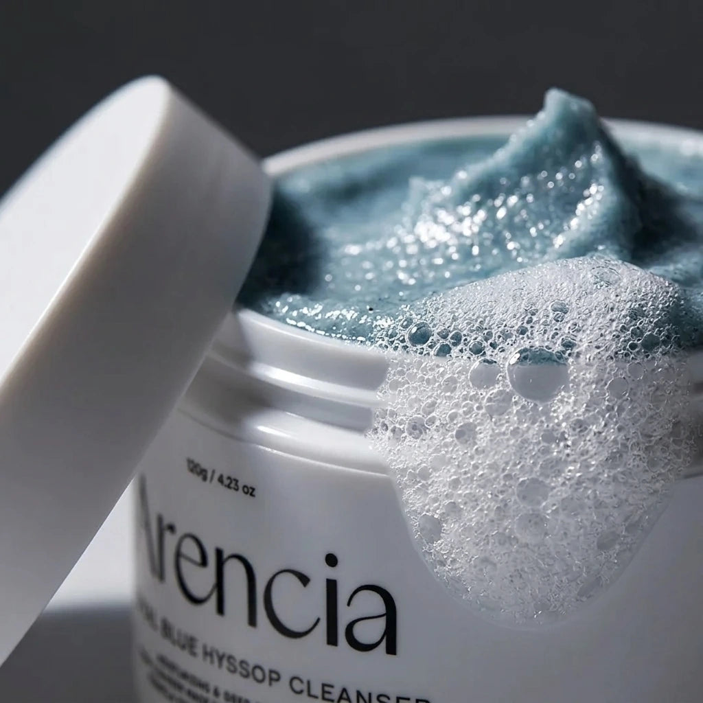 Angle : Gros plan macro très serré, focus sur la mousse et le plastique du pot.  Scène : Un gros plan extrême sur la texture mousseuse blanche qui déborde sur le côté droit du pot d'Arencia. On distingue parfaitement les micro-bulles d'air transparentes de la mousse. La texture bleue sculptée est visible au second plan. Le couvercle blanc mat est posé de biais, légèrement flou à gauche.  Fond & Lumière : Arrière-plan minimaliste sombre. Un éclairage rasant fait miroiter la surface humide du nettoyant bleu, 