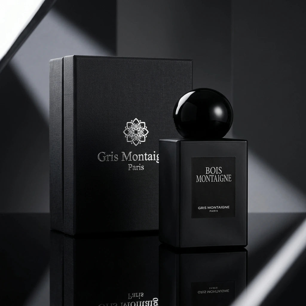 Angle : Prise de vue frontale, mettant en avant le design et le graphisme de la marque.  Fond : Un arrière-plan minimaliste et abstrait aux nuances de gris anthracite et noir mat.  Mise en scène : Une luxueuse boîte ou un coffret noir fermé "Gris Montaigne Paris" avec son logo floral argenté est placé à l'arrière. Juste devant, légèrement excentré, le flacon de parfum "Bois Montaigne" est posé, créant un reflet subtil sur un sol miroir noir brillant.  Lumière : Éclairage publicitaire de studio, faisceaux lu