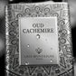 Un plan macro très serré concentré sur l'étiquette centrale rectangulaire 'OUD CACHEMIRE' sur le flacon de image_1fe27b.jpg. De fines micro-gouttelettes d'eau pure perlent de manière réaliste sur la surface externe argentée et sur les lignes complexes du mandala. L'arrière-plan présente un flou artistique (bokeh) très doux composé de nuances gris perle et noires. Style : Photographie de produit sensorielle et haut de gamme. (gemini-3.1-flash-image-preview)