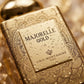 Un plan macro très serré concentré sur la plaque centrale rectangulaire 'MAJORELLE GOLD' sur le flacon de image_2046de.jpg. De fines micro-gouttelettes d'eau pure perlent de manière réaliste sur la surface externe dorée et sur les lignes complexes du mandala. L'arrière-plan présente un flou artistique (bokeh) très doux aux nuances chaudes et dorées. Style : Photographie de produit sensorielle et haut de gamme. (gemini-3.1-flash-image-preview)