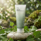 E-commerce lifestyle shot of the Anua Heartleaf Quercetinol Pore Deep Cleansing 150ml tube (image_0.png). The product stands on a damp, smooth light grey river stone. It is surrounded by fresh green Houttuynia cordata (heartleaf) leaves and a few scattered droplets of clear water, suggesting a pore-deep clean. The background is a gently blurred natural setting with hints of sunlight filtering through leaves. Dimensions: 1200x1200px. Fresh, organic, and clean aesthetic. (gemini-3.1-flash-image-preview)
