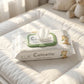 Commercial 1:1 product photography of "Calinette - Lingettes Bébé Sensitive" packaging placed on a white padded changing mat. Gentle, warm sunbeams stream in from a nearby window, creating a bright, pure, and realistic atmosphere. One wipe is slightly peeking out of the closed plastic lid. Crisp details. Shot on 70mm lens, f/4.0, ISO 250, realistic textures, no 3D render. --ar 1:1 (gemini-3.1-flash-image-preview)