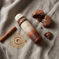 Angle : Nadir parfait (Zénithal à 90 degrés).  Scène : Le flacon de l'image image_8bb442.jpg est allongé horizontalement. Autour de lui, la matière s'exprime : une coque de macaron au chocolat brisée laissant échapper sa ganache, un bâton de cannelle brut et une traînée géométrique parfaite de caramel liquide doré dessinant une spirale sur le sol.  Fond & Éclairage : Une nappe en lin lourd, froissée et texturée, couleur grège (entre le beige et le gris). Éclairage latéral très doux imitant une lumière natur
