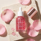 Angle : Flat Lay serré axé sur le flacon.  Scène : Le flacon de l'image image_cd16ff.png est entouré de pétales de rose rose tendre parsemés de fines gouttes d'eau. Un couvercle métallique or rose brillant est posé de manière asymétrique sur le côté pour apporter une touche de brillance. Quelques gouttes de gel rose transparent sont disposées sur le support à côté de la bouteille.  Fond & Éclairage : Une dalle en pierre de travertin crème très texturée. Lumière rasante douce pour mettre en relief les contou