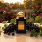 Un plan large d'un flacon de image_2.png posé sur une table en travertin poli, sur une terrasse donnant sur un jardin de roses anciennes à la tombée de la nuit. La lumière est chaude et dorée (golden hour). Autour de image_2.png, des grappes de raisin de Corinthe et des feuilles de géranium créent un cadre organique. La plaque dorée de image_2.png reflète l'heure dorée. Style : Photographie éditoriale de luxe, sensuelle. (gemini-3-pro-image-preview)