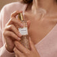 Un plan serré et intimiste centré sur les mains d'un mannequin femme à la peau satinée et impeccable. D'une main, elle tient délicatement le corps cylindrique du flacon de image_16.png et en presse le spray, libérant un nuage fin, vaporeux et aérien de brume parfumée vers son cou. Le fond montre la texture floue d'un pull en cachemire rose pâle très doux. Style : Campagne publicitaire de beauté et de soin de la peau. (gemini-3-pro-image-preview)