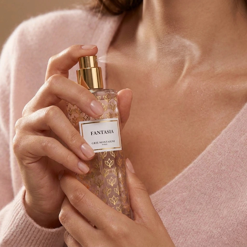 Un plan serré et intimiste centré sur les mains d'un mannequin femme à la peau satinée et impeccable. D'une main, elle tient délicatement le corps cylindrique du flacon de image_16.png et en presse le spray, libérant un nuage fin, vaporeux et aérien de brume parfumée vers son cou. Le fond montre la texture floue d'un pull en cachemire rose pâle très doux. Style : Campagne publicitaire de beauté et de soin de la peau. (gemini-3-pro-image-preview)