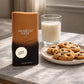 Un visuel lifestyle chaleureux à 1200x1200px. Le parfum d'image_93c92b.jpg est installé sur une table basse en marbre blanc. À côté du flacon, une assiette en céramique minimaliste présente des cookies artisanaux encore chauds avec des pépites fondantes, accompagnés d'un verre de lait. Une lumière naturelle traverse une fenêtre invisible, projetant des ombres douces et réalistes sur la scène. (gpt-image-1.5)