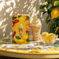 Un visuel lifestyle ensoleillé à 1200x1200px. Le parfum de image_93d566.jpg est posé sur une table en céramique italienne peinte à la main. À côté du flacon, une coupelle transparente contient deux boules de sorbet au citron frais avec une cuillère dorée. Une lumière naturelle vive traverse des feuillages invisibles, projetant des ombres douces et réalistes de feuilles de citronnier sur la scène. (gpt-image-2)