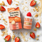 Une vue de dessus (flat-lay) artistique et lumineuse sur un format carré de 1200x1200px. Au centre, le flacon de parfum en forme de gâteau à étages et sa boîte "Arabiyat Sugar - Strawberry Tres Leches" de image_935887.jpg l'un à côté de l'autre. Ils sont entourés de morceaux de fraises rouges fraîches et juteuses, de fines éclaboussures de lait concentré sucré et d'une légère pluie de miettes de génoise dorée. L'ensemble est disposé sur une table en marbre blanc brillant sous une lumière studio douce et env