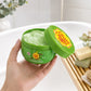 A commercial frontal photograph (angle frontal) in a bright bathroom. A woman's hand, with manicured nails, holds the 200ml Chupa Chups - Apple Crush - Whipped Soap pot. The yellow lid is partially unscrewed and tilted, revealing the fluffy, green-swirled whipped soap inside. The background shows a white bathtub with some unbranded artisan soaps on a wooden rack. Light reflects on the product label. (gpt-image-2)